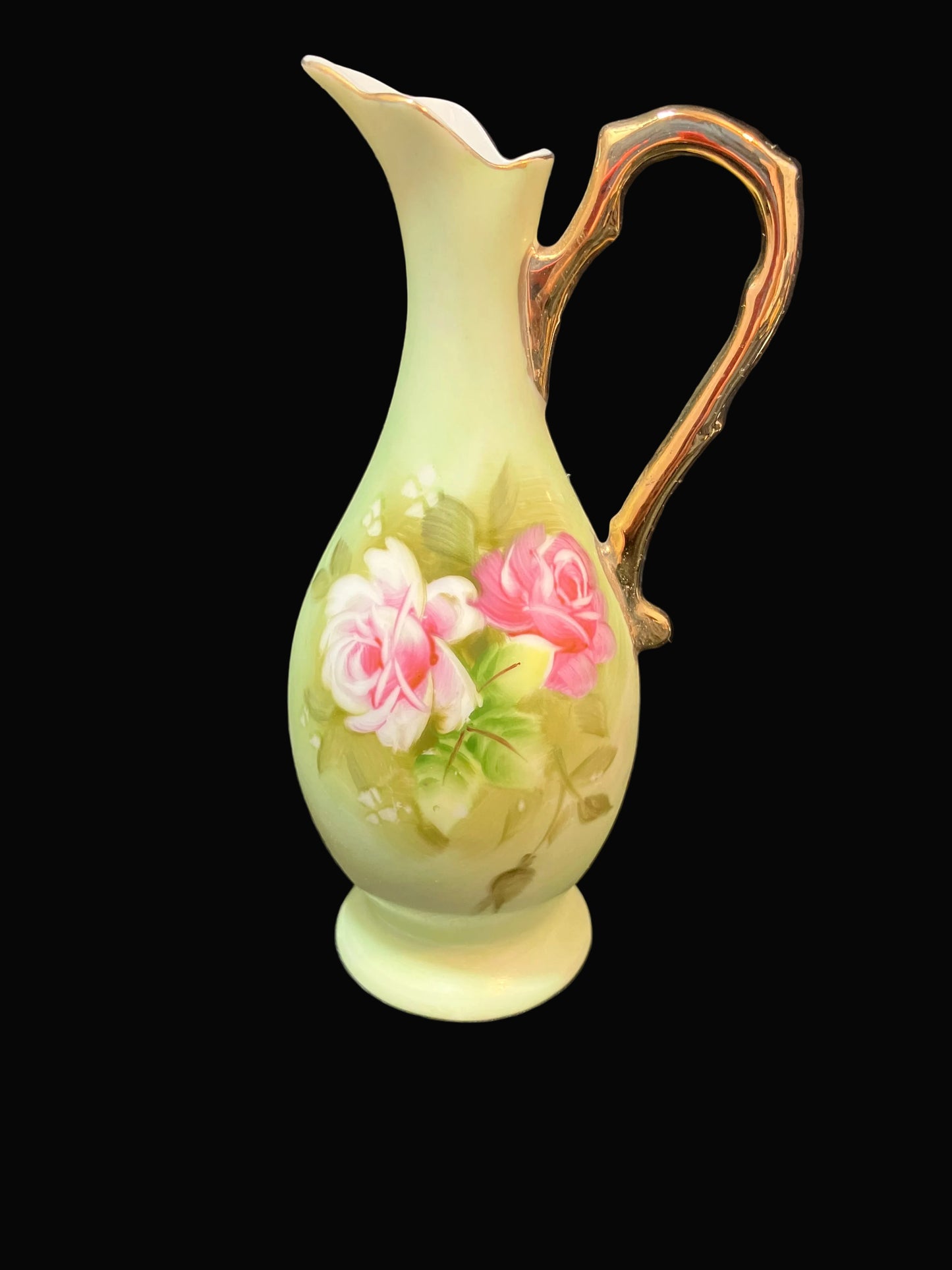 A vintage mini Lefton Japan ceramic pitcher with a pale yellow-green body, hand-painted pink and white roses with green leaves, and a metallic gold handle, set against a solid black background.