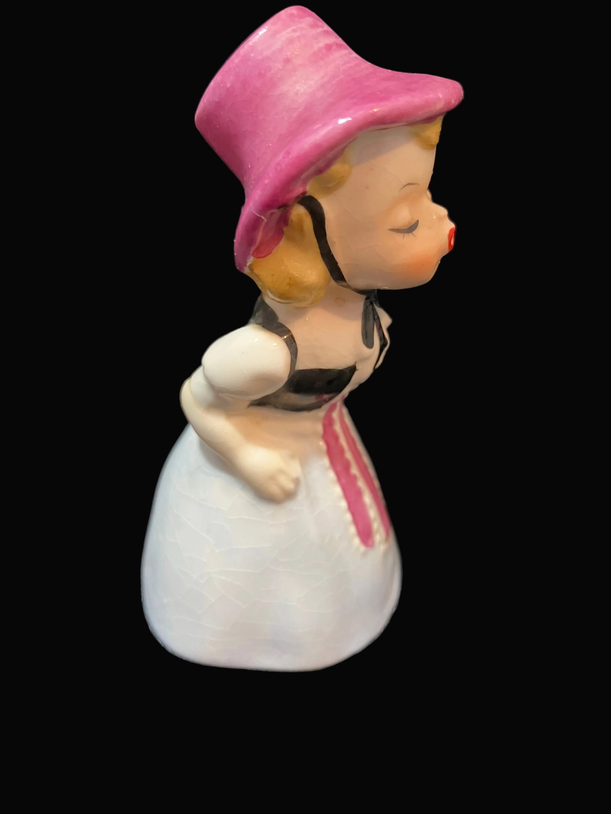 A vintage ceramic figurine of a young girl in profile, wearing a pink bonnet and a white dress with pink and black details, set against a solid black background.