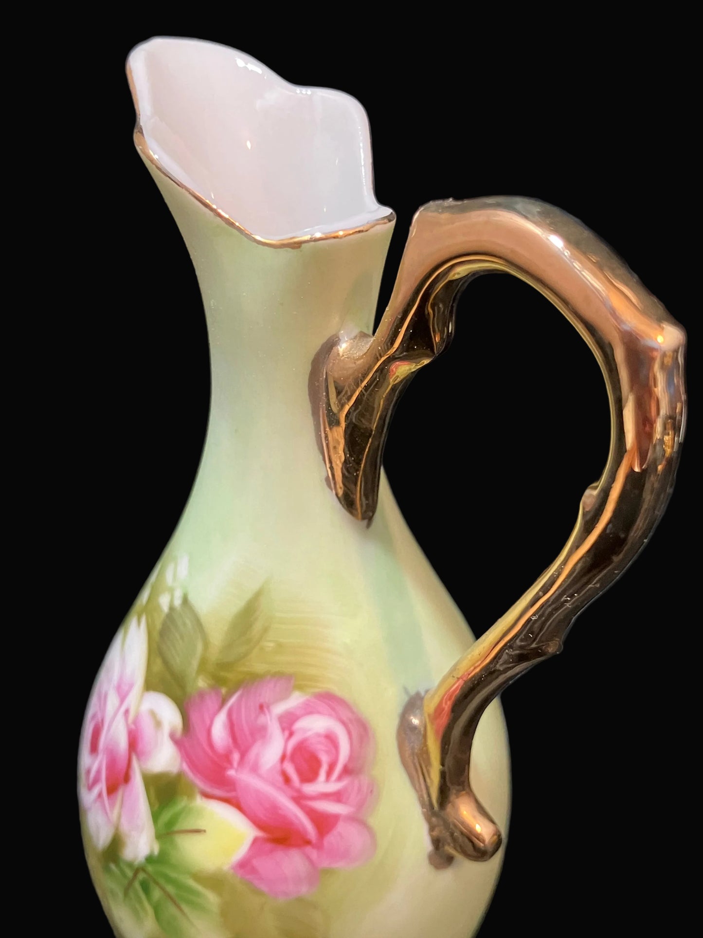 A vintage-style ceramic pitcher with a pale yellow-green body, a hand-painted pink rose and green leaves on the side, and a prominent curved gold-tone handle, set against a dark textured fabric background.