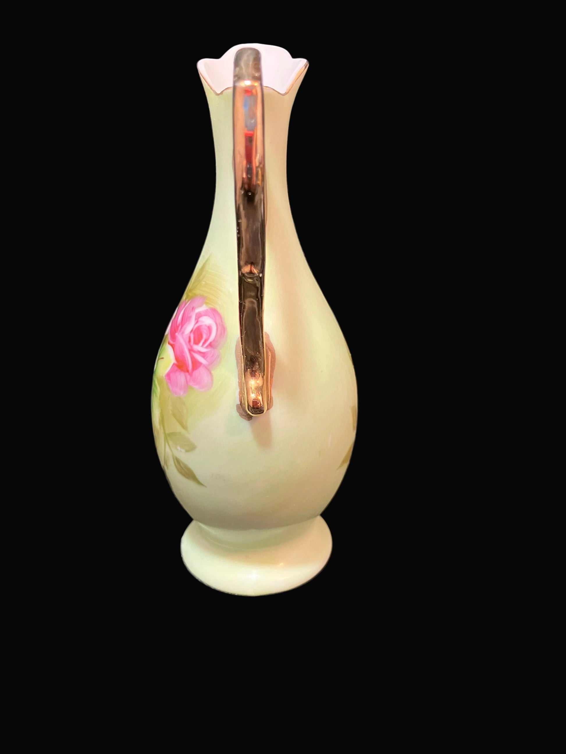A vintage soft green-colored ceramic pitcher with a flared neck, a vertical handle, and a pink rose decoration, shown against a solid black background.