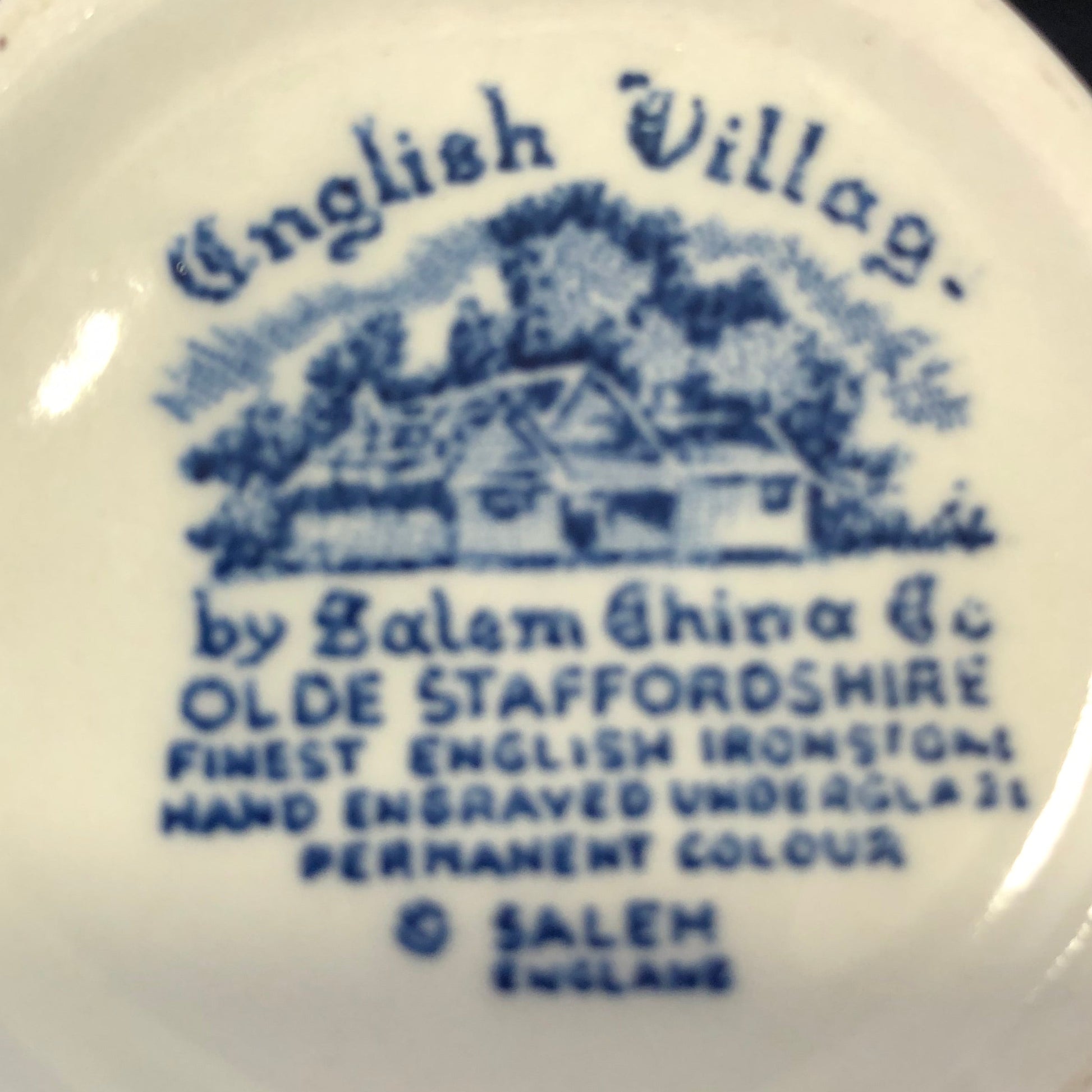 Close-up of the underside of a ceramic creamer held in a hand, showing blue printed text reading 'English Village.' and 'by Salem China Co Olde Staffordshire' with a blue house illustration on an off-white porcelain surface.