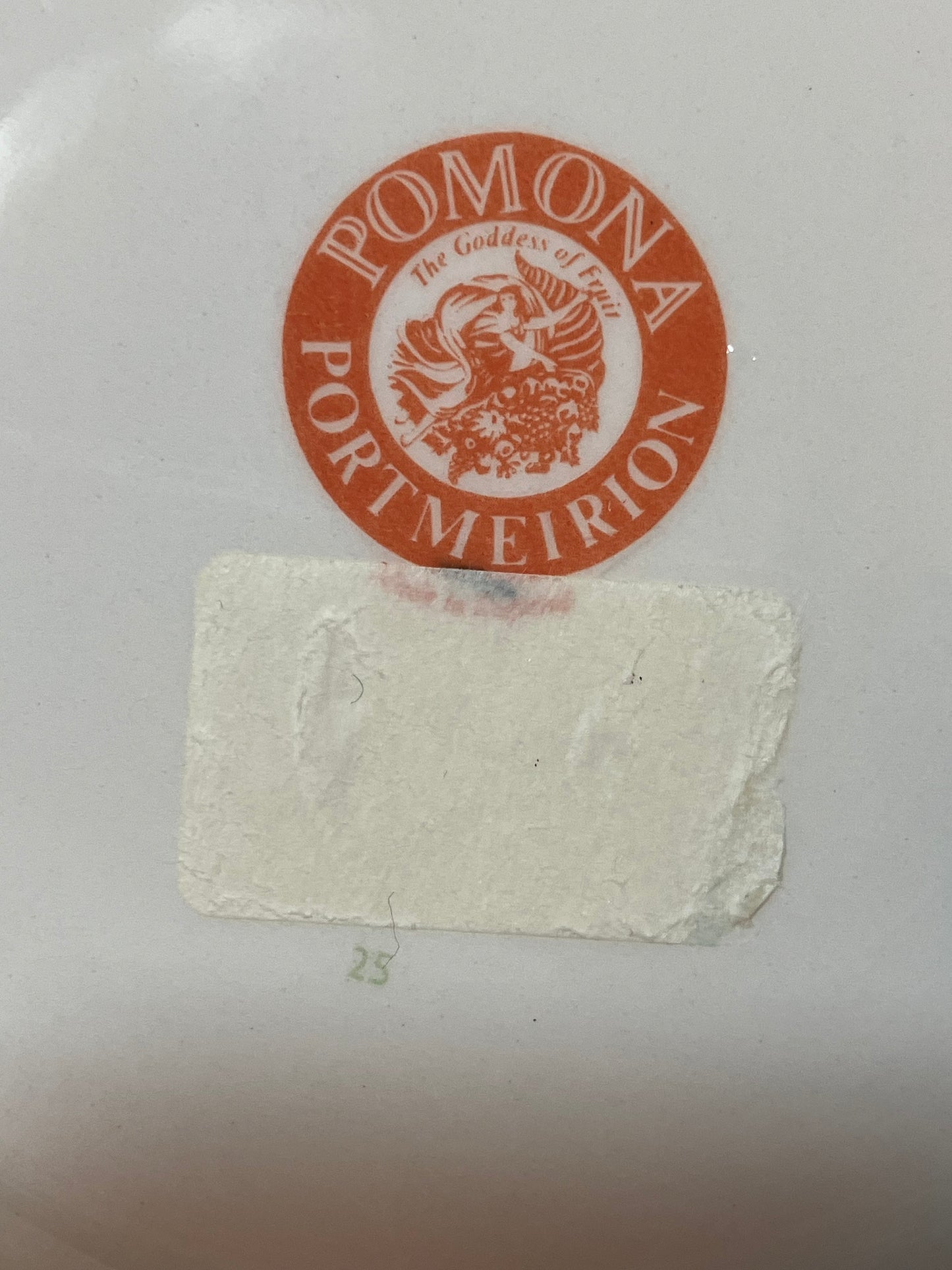 A close-up of a white surface featuring a circular orange and white sticker that reads 'POMONA PORT MEIRION' with a central figure emblem, and a rectangular off-white sticker applied below it.