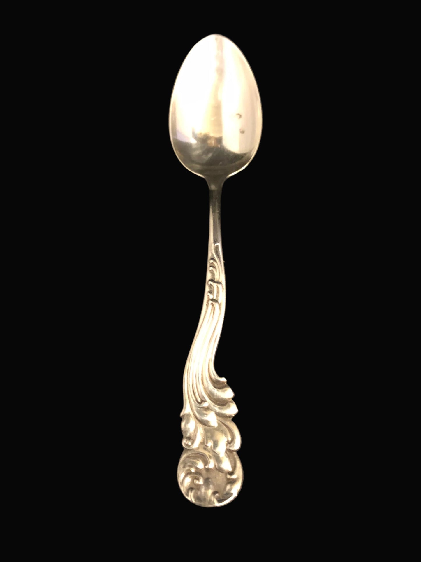 A vintage sterling silver spoon with an ornate, decorative handle, shown against a dark black background.