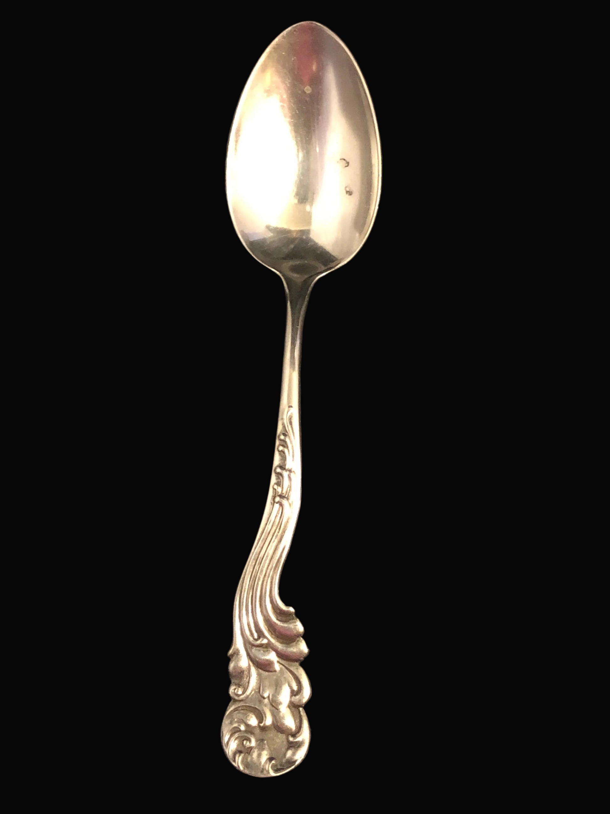 A vintage sterling silver spoon with an ornate, decorative handle, shown vertically against a dark background.