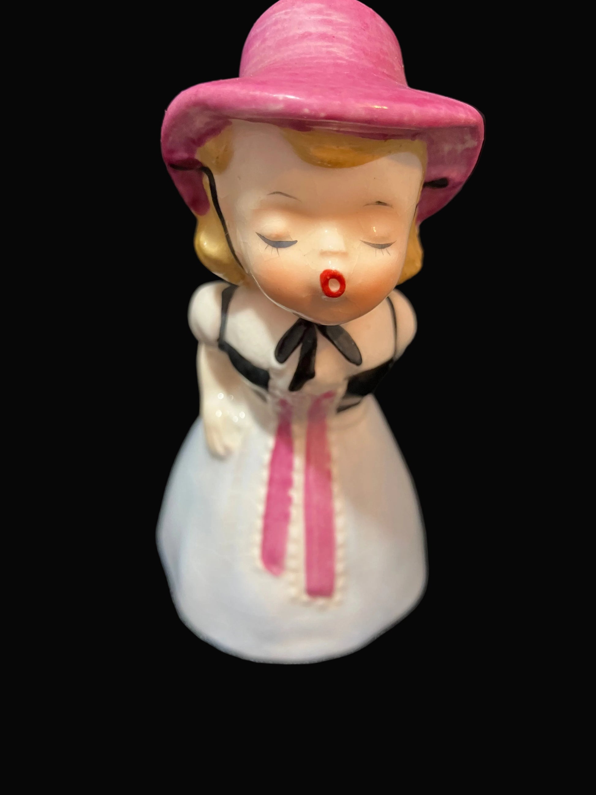 A vintage ceramic figurine of a girl wearing a pink hat and a white dress with a pink ribbon and black bow, set against a solid black background.