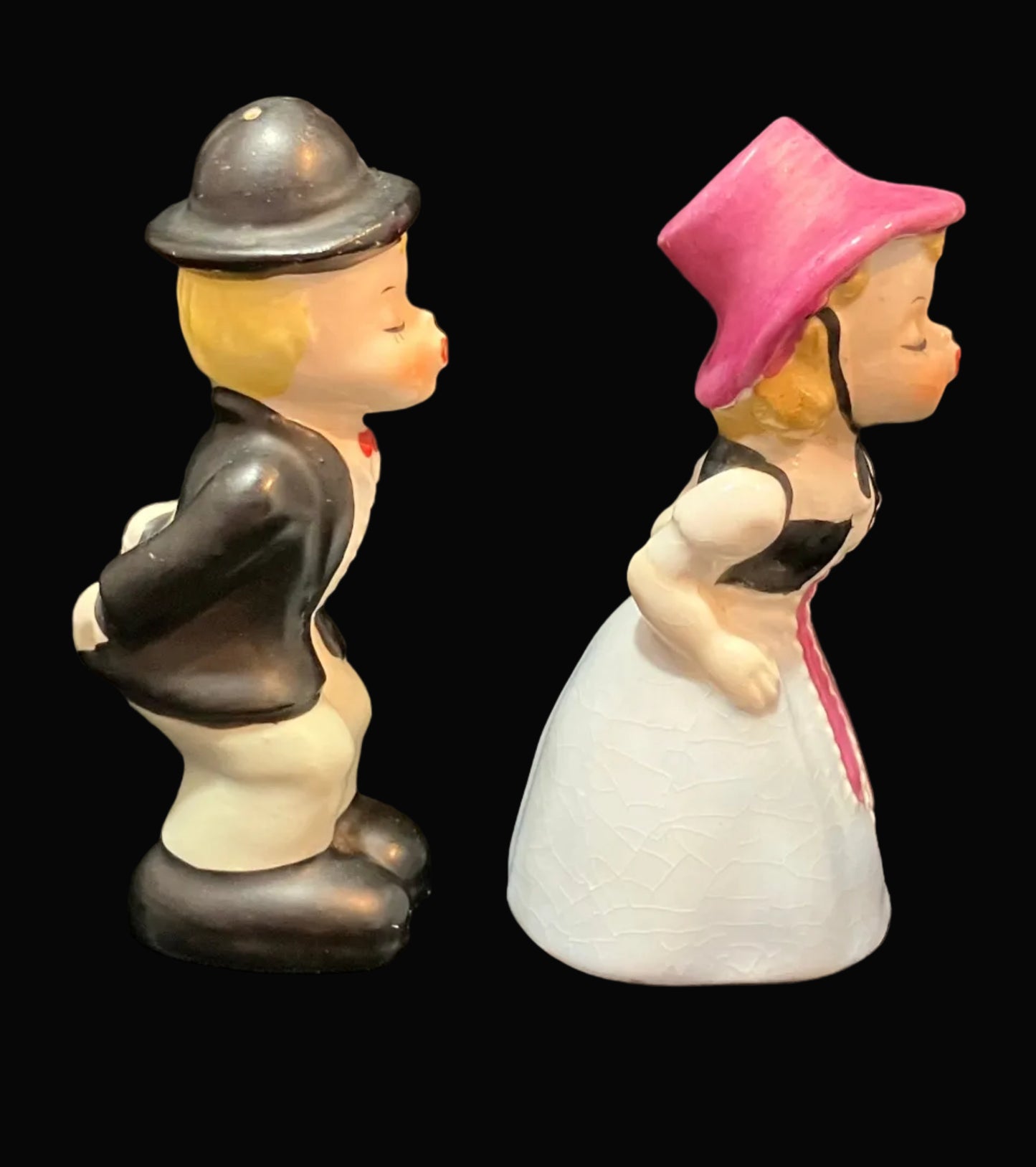 A pair of vintage ceramic salt and pepper shakers depicting a man and a woman in profile, set against a solid black background. The man wears a black jacket, white pants, and a black bowler hat.