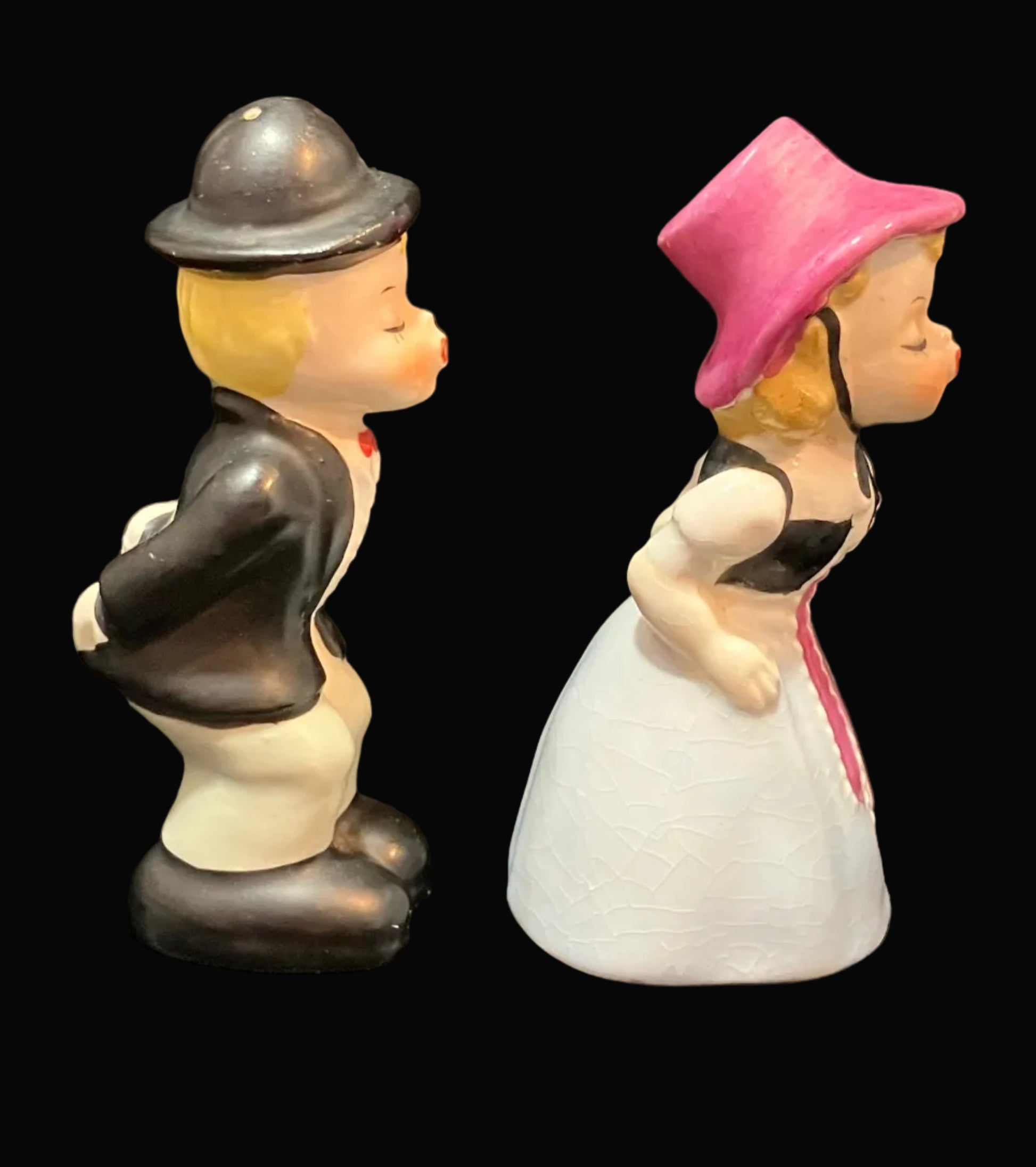 A pair of vintage ceramic salt and pepper shakers depicting a man and a woman in profile, set against a solid black background. The man wears a black jacket, white pants, and a black bowler hat.