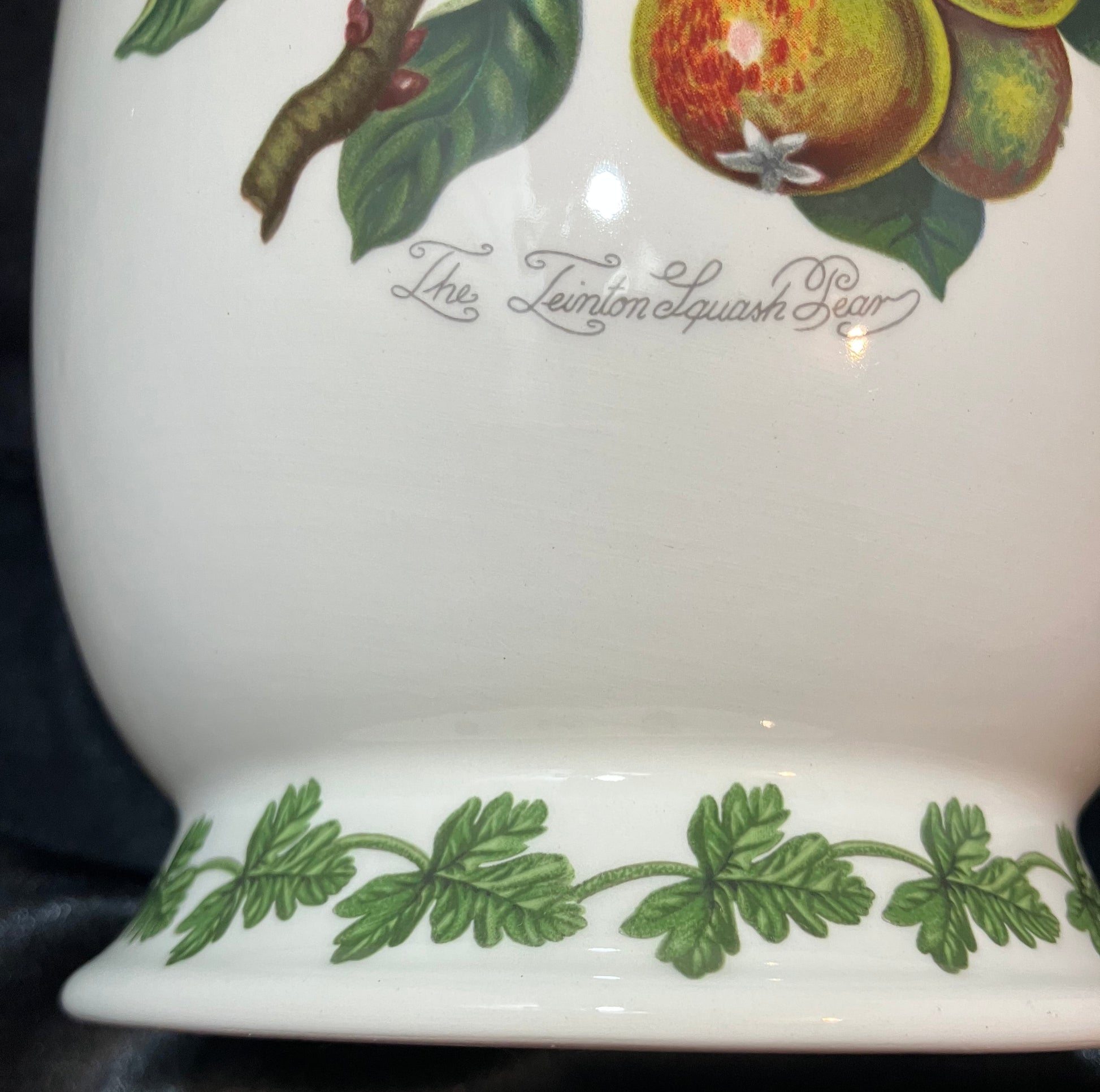 Close-up of a white ceramic container or vase with a green leaf border and fruit illustration near the top, with cursive text reading 'The ternion squash pear' on a dark fabric background.