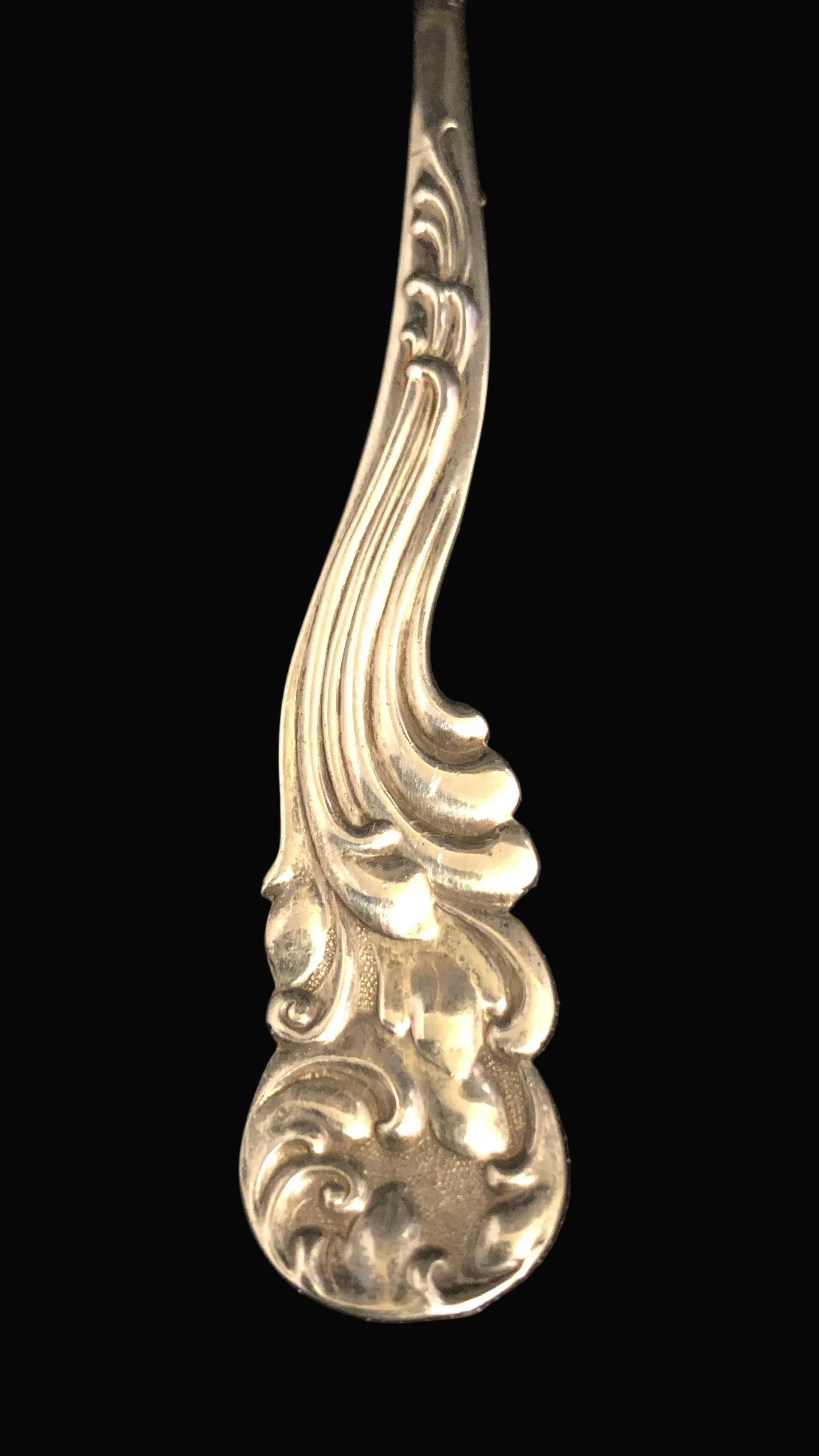 A close-up of a polished silver spoon handle with an ornate, decorative scrollwork pattern