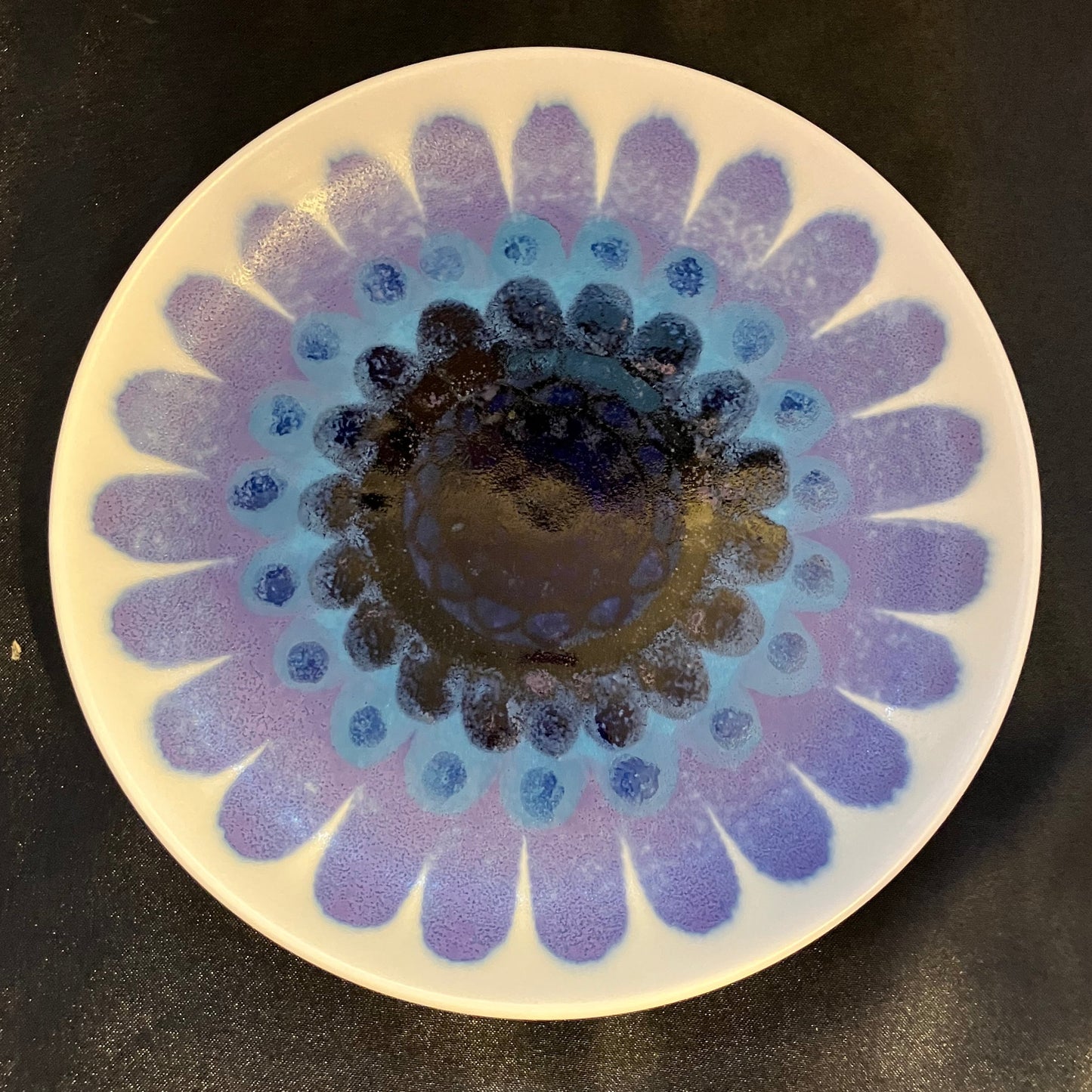 A round ceramic plate with a hand-painted daisy-like floral pattern in shades of blue, purple, and black on a white base, photographed from above on a dark surface.