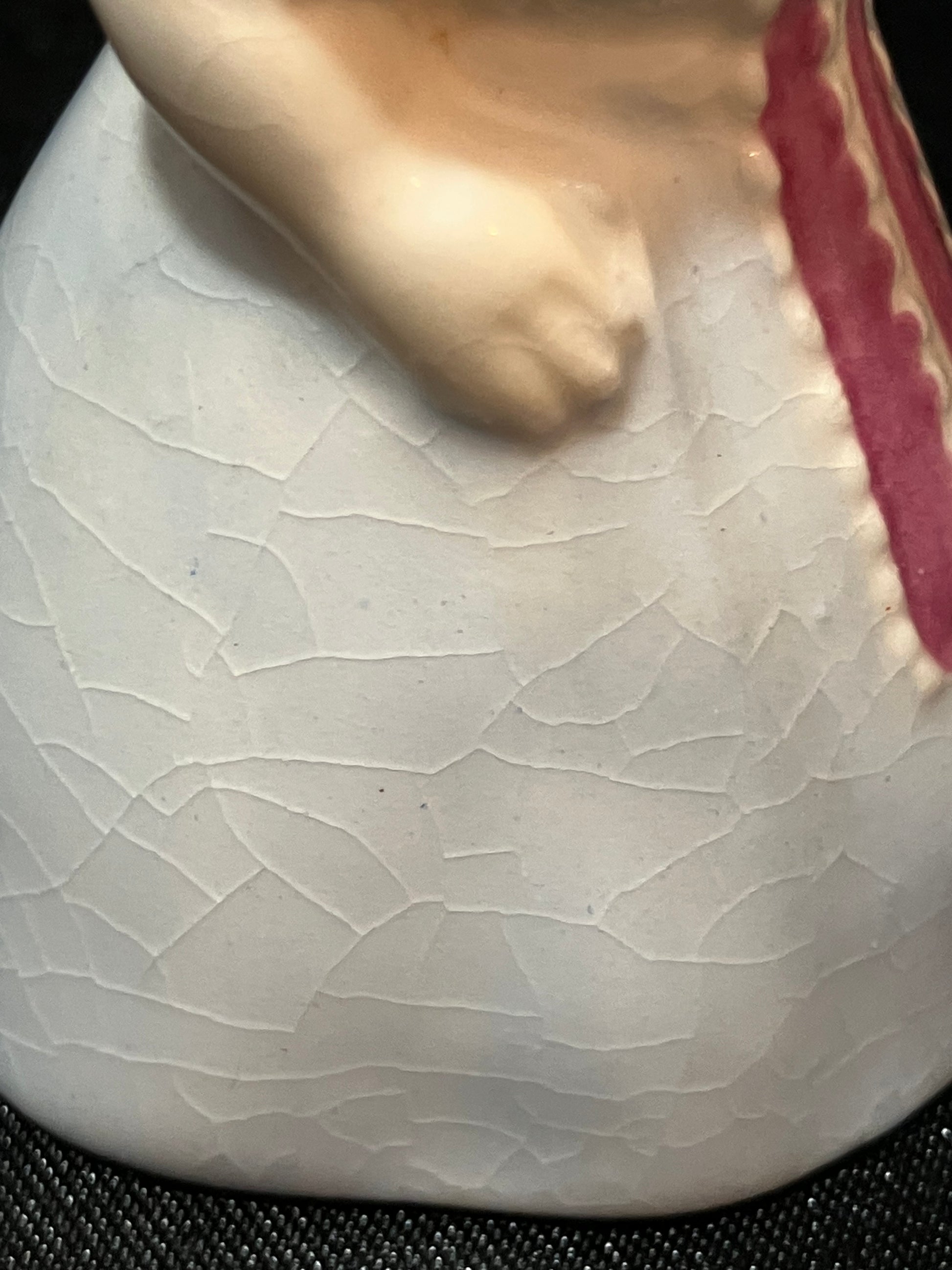 A close-up of a ceramic plate with a white glaze featuring a crackle pattern, a pink scalloped rim, and a beige sculpted hand emerging from the left side, set against a black textured background.