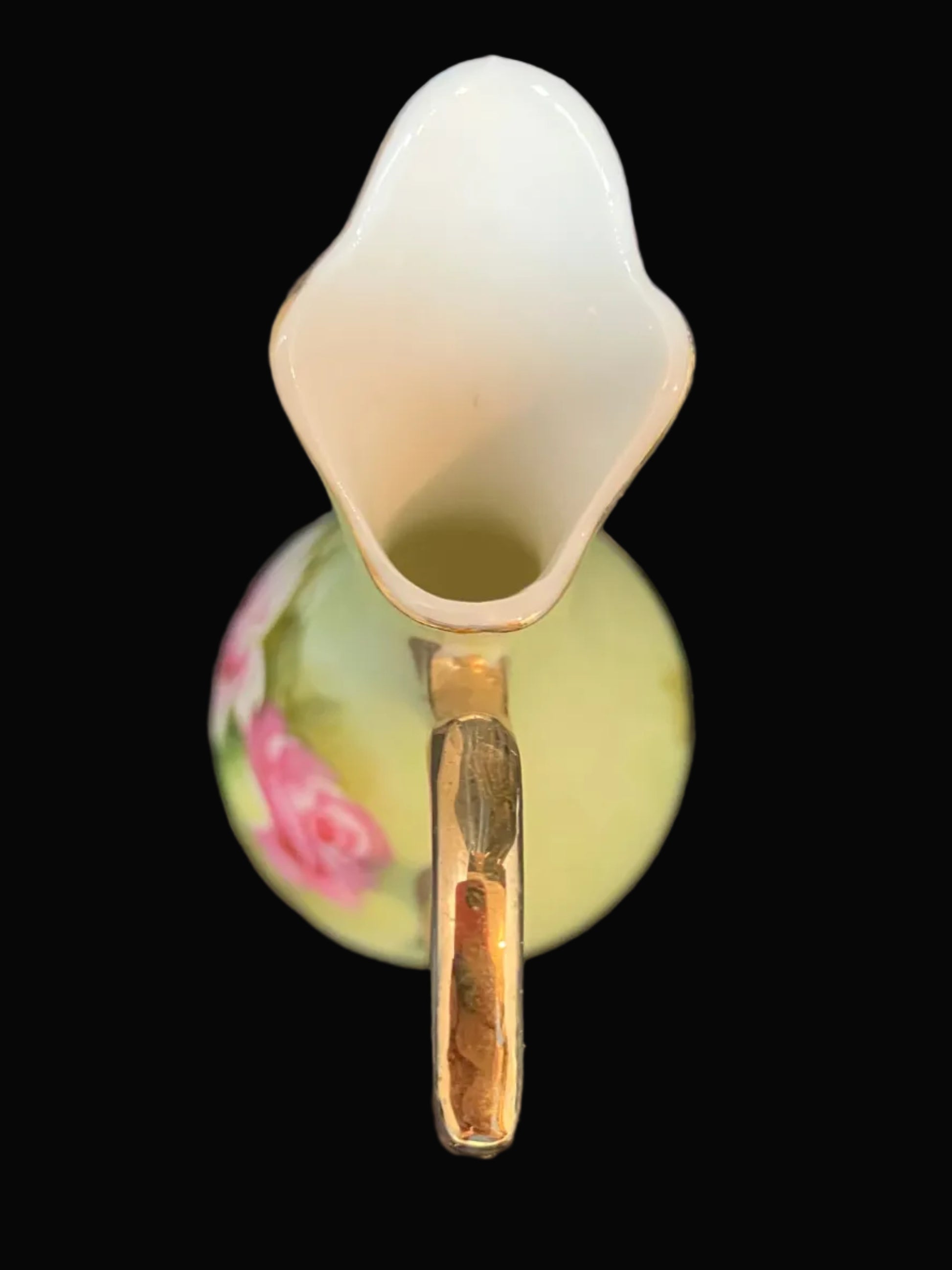 A vintage ceramic pitcher viewed from above, featuring a glossy white flared spout, a gold-toned handle, and a pale yellow-green body decorated with pink floral patterns, set against a solid black background.