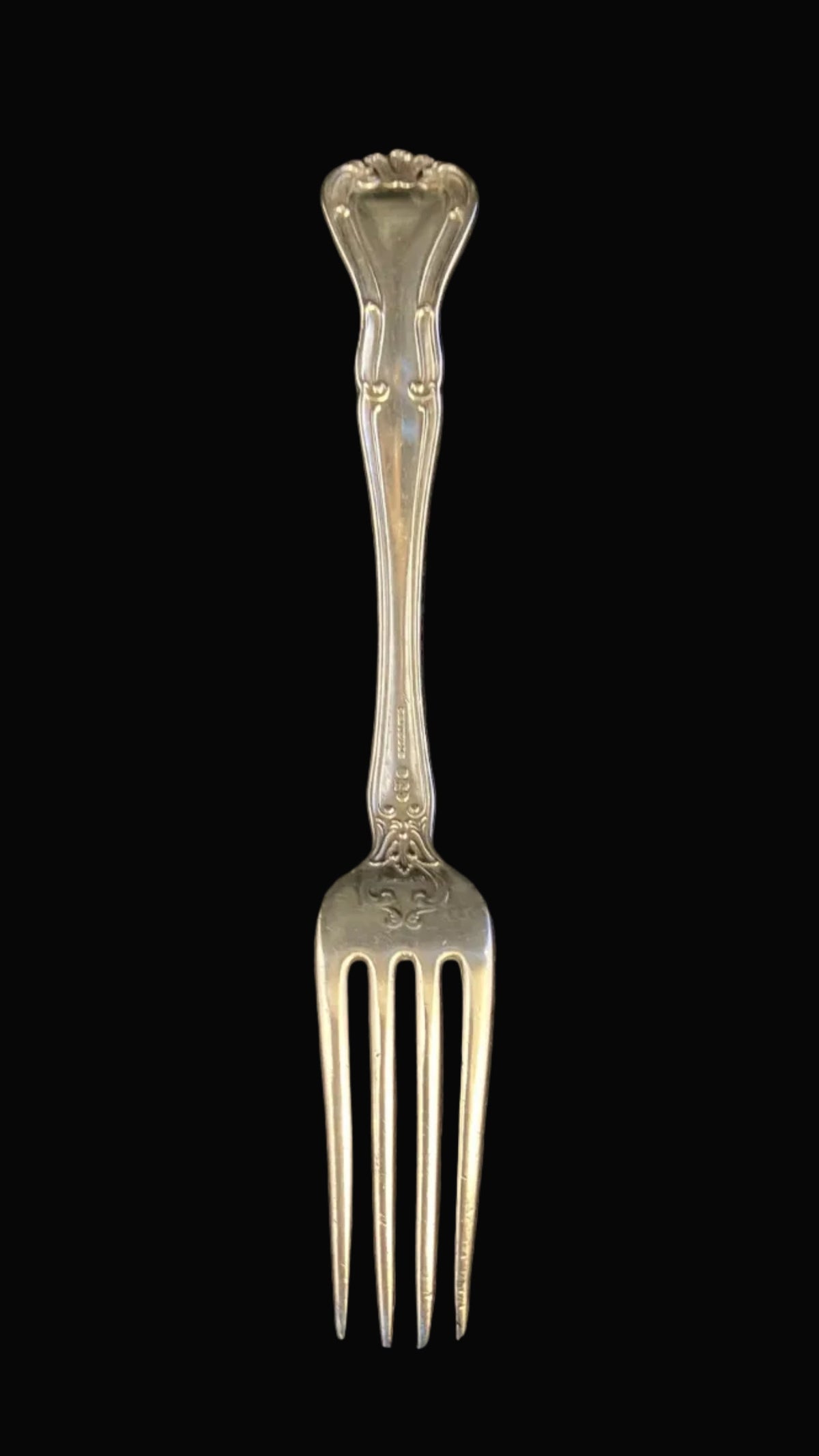 An ornate vintage sterling silver serving fork with a decorative handle, displayed vertically on a dark textured fabric background.