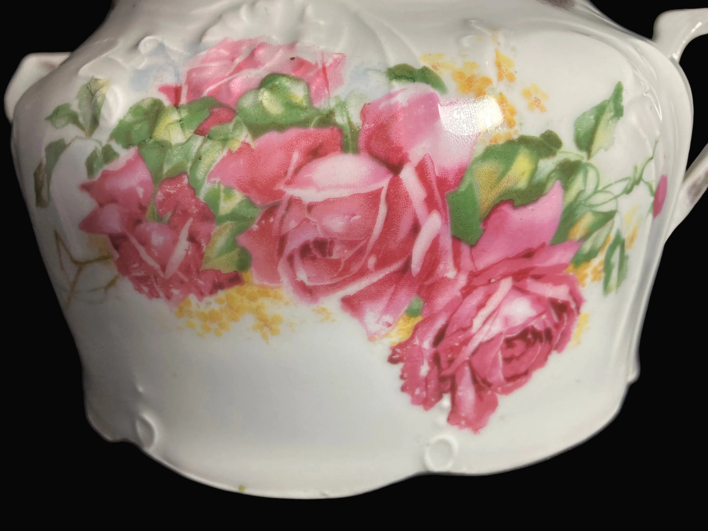A close-up of a white ceramic tureen or lidded dish decorated with a hand-painted floral design of pink roses and green leaves, resting on a blacksurface.
