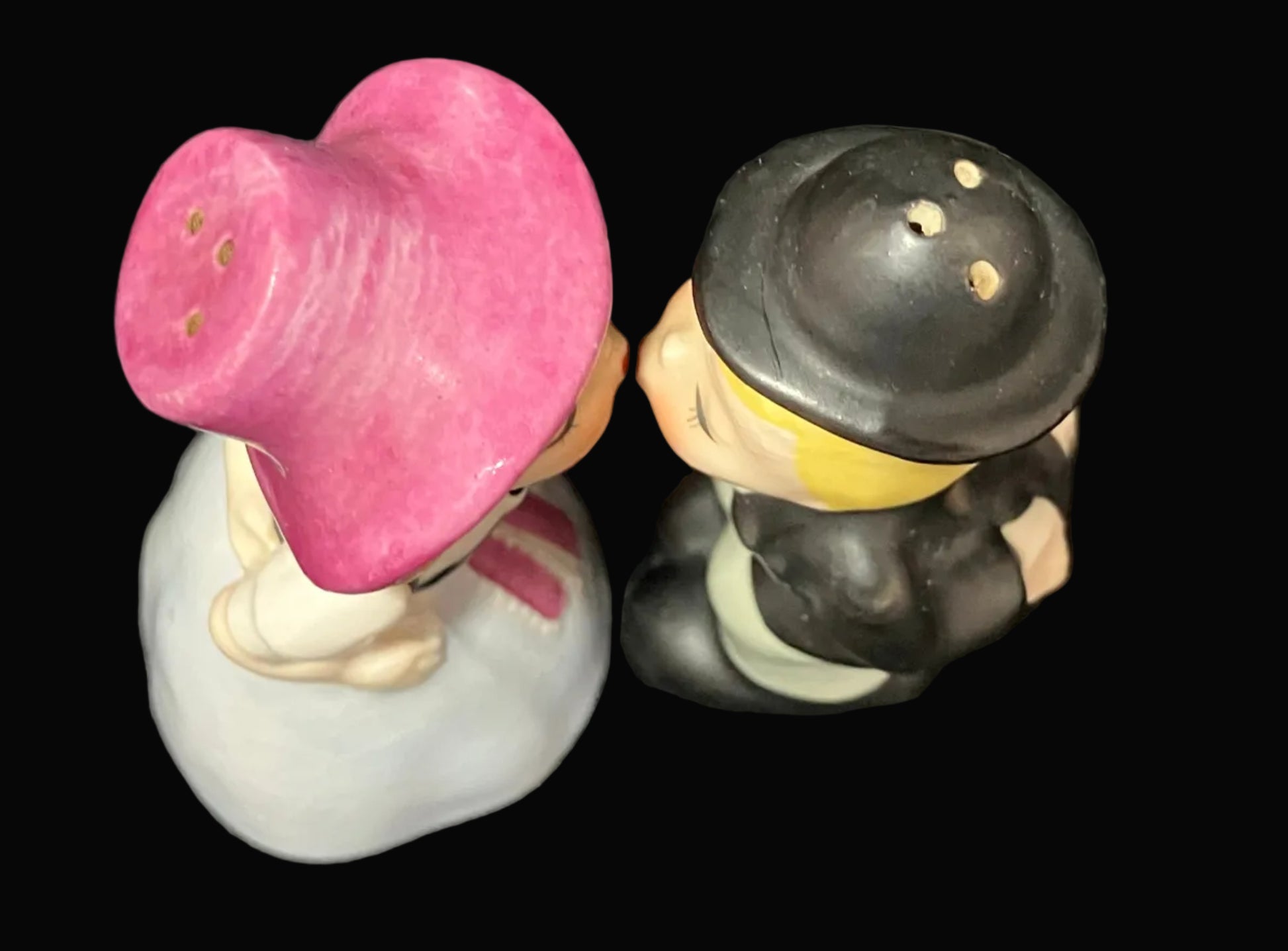 A pair of ceramic salt and pepper shakers shaped like a couple embracing, photographed against a solid black background. The woman figure wears a pink hat and white dress, and the man figure wears a black hat and black jacket.