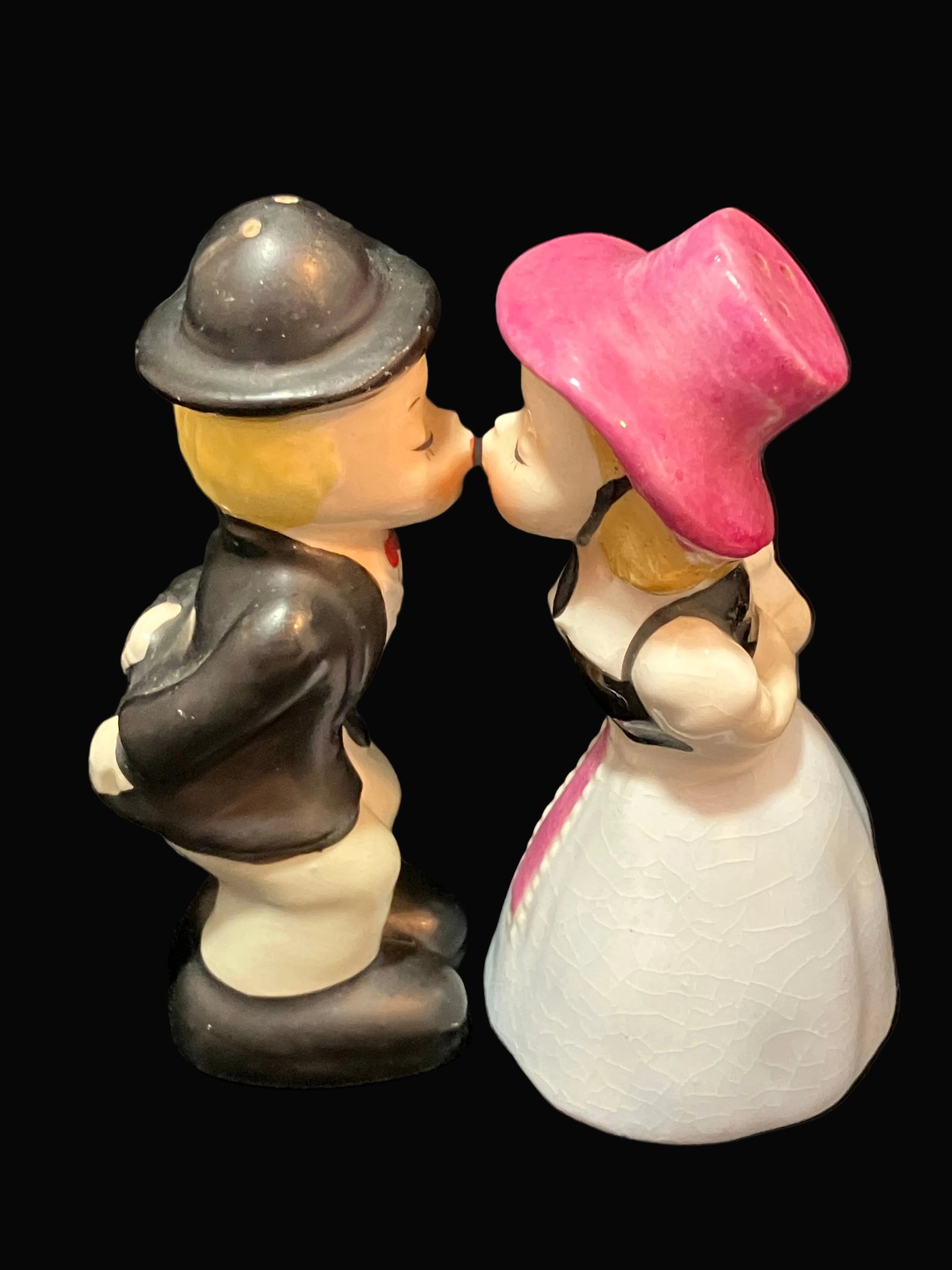 A vintage ceramic salt and pepper shaker figurine depicting a kissing couple. The man wears a black bowler hat, black jacket, and cream trousers, while the woman wears a bright pink top hat and a white dress with a black bodice.