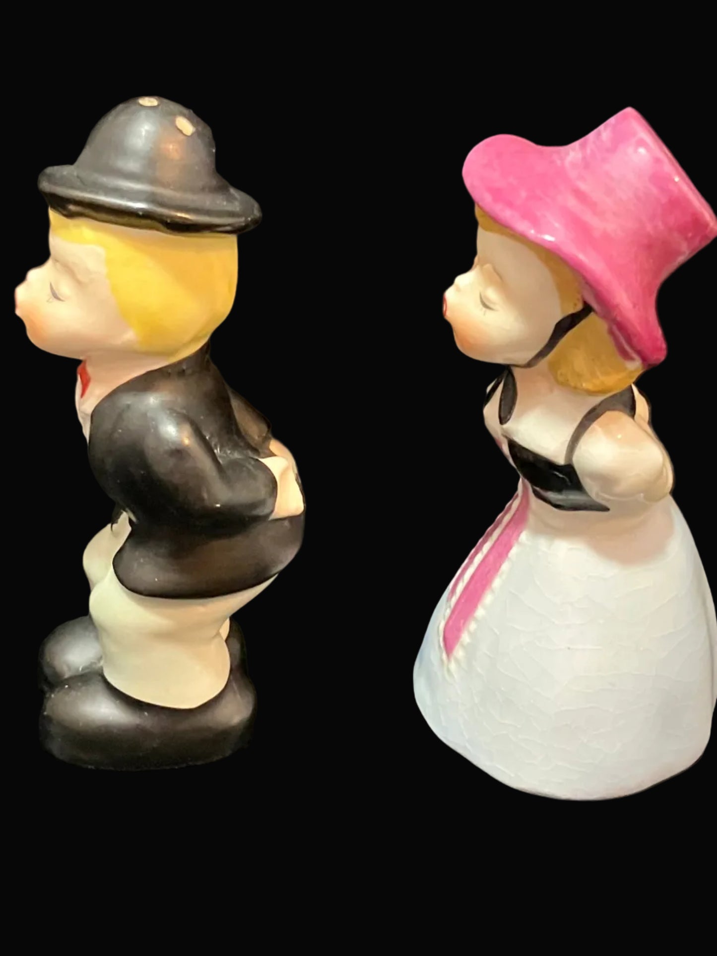 A pair of vintage ceramic figurines depicting a couple in profile, set against a solid black background. The male figure wears a black hat and jacket with cream pants and yellow hair.