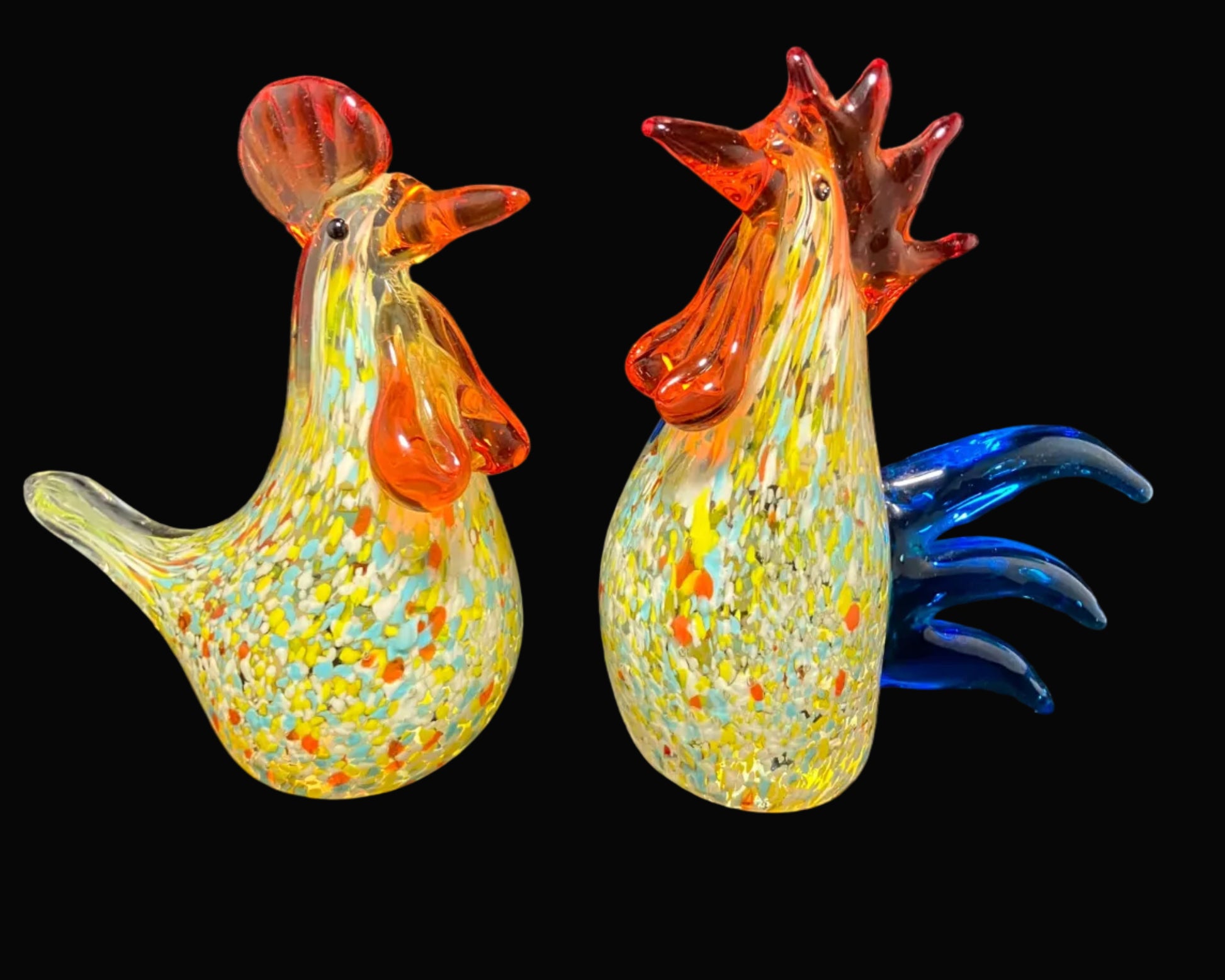 Two decorative hand-blown glass rooster figurines on a dark gray fabric backdrop. One rooster has orange-red comb and wattle with yellow, green, and orange speckles, and blue tail feathers.