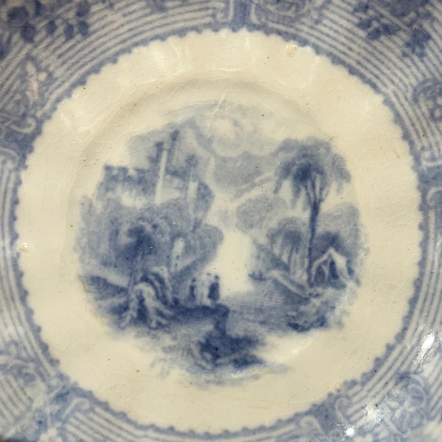 Close-up of the inside of a blue and white porcelain saucer, showing a central landscape illustration surrounded by decorative floral and geometric borders.