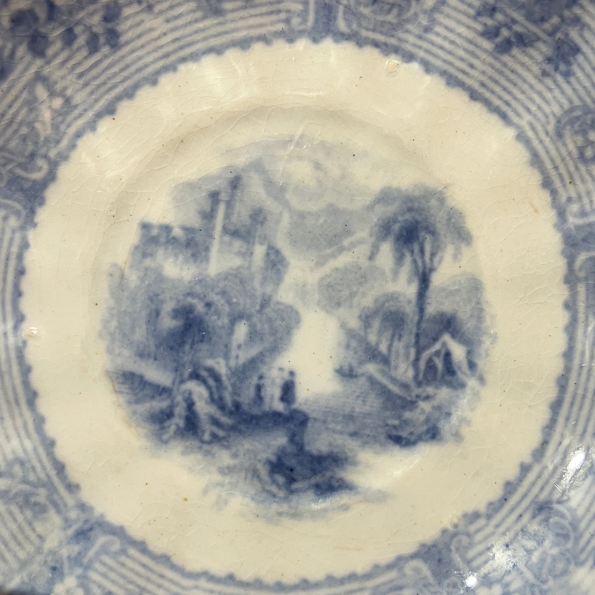 Close-up of the inside of a blue and white porcelain saucer, showing a central landscape illustration surrounded by decorative floral and geometric borders.