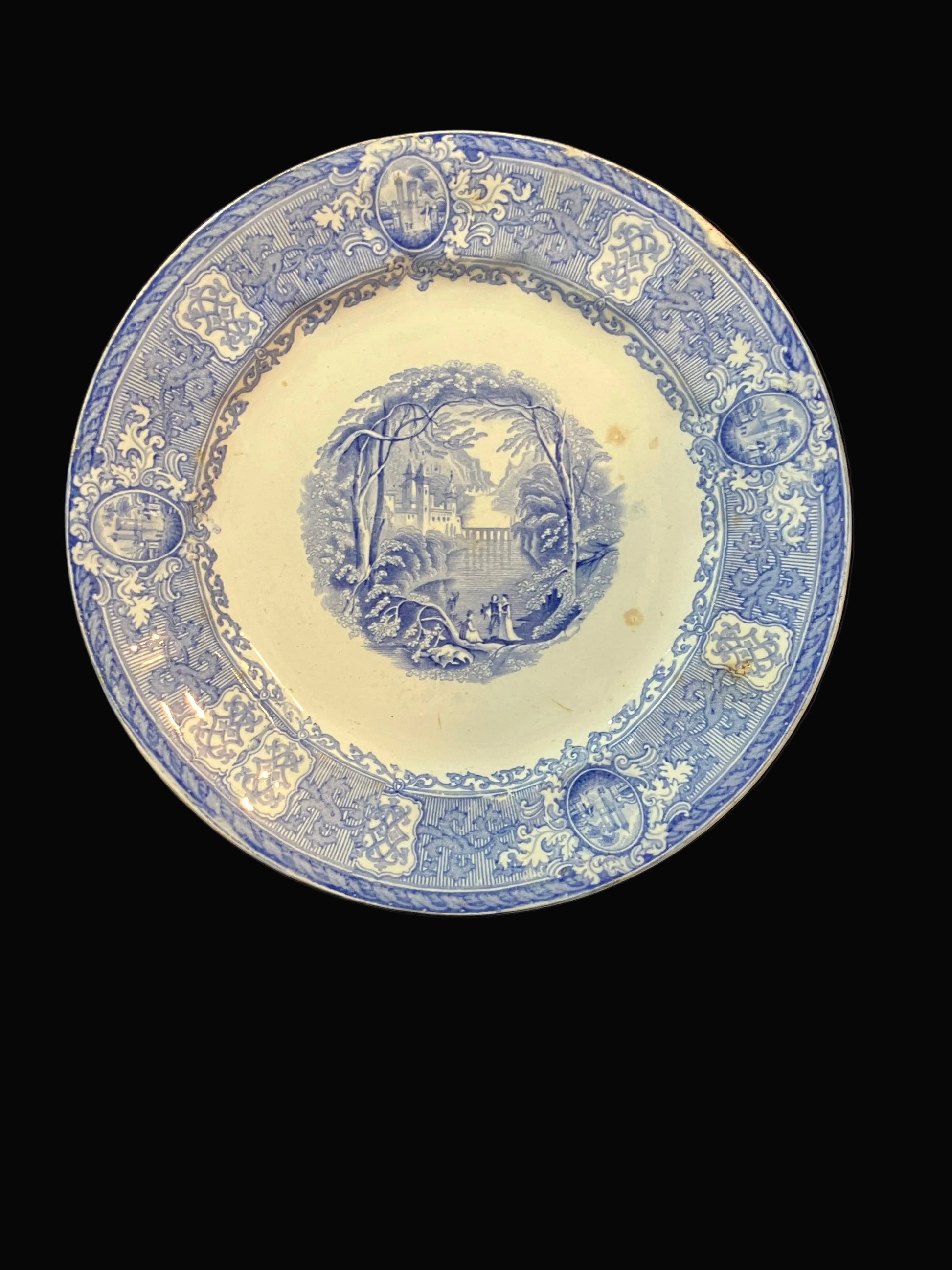 A vintage blue and white ceramic plate with an illustrated landscape design in the center and decorative border motifs, displayed against a black background.