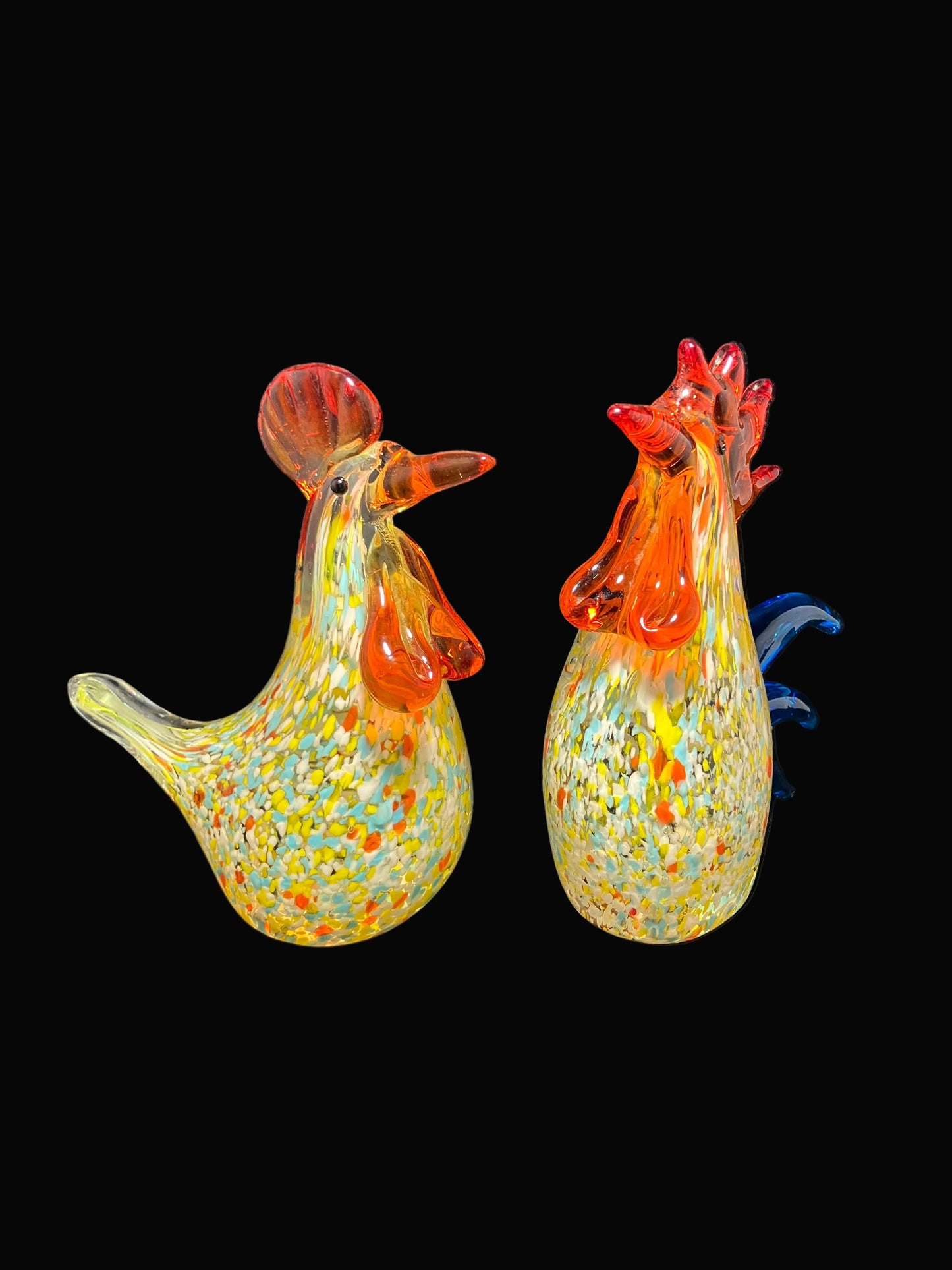 Two hand-blown glass rooster figurines with orange-red combs and wattle heads, speckled with yellow, green, and blue glass.