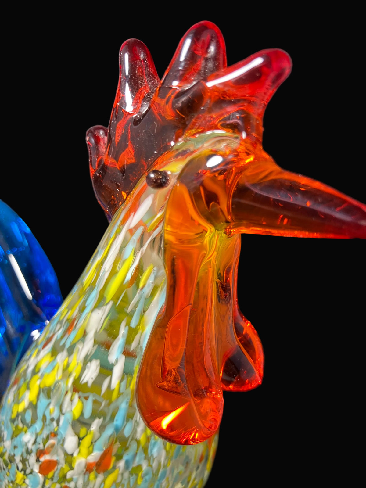 A close-up of a hand-blown glass rooster figurine with a translucent red and orange comb and beak, and a multicolored speckled body in yellow, blue, green, and white, set against a dark background.