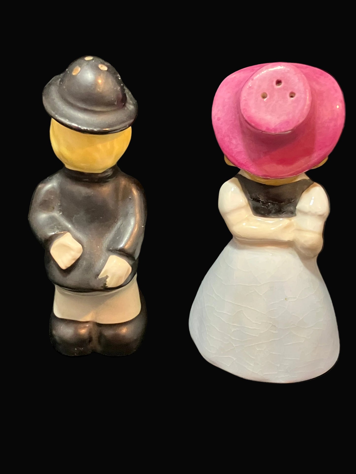 Two small ceramic figurines on a black background, one with a dark gray hat and jacket, and cream pants, and the other with a pink hat and white dress.