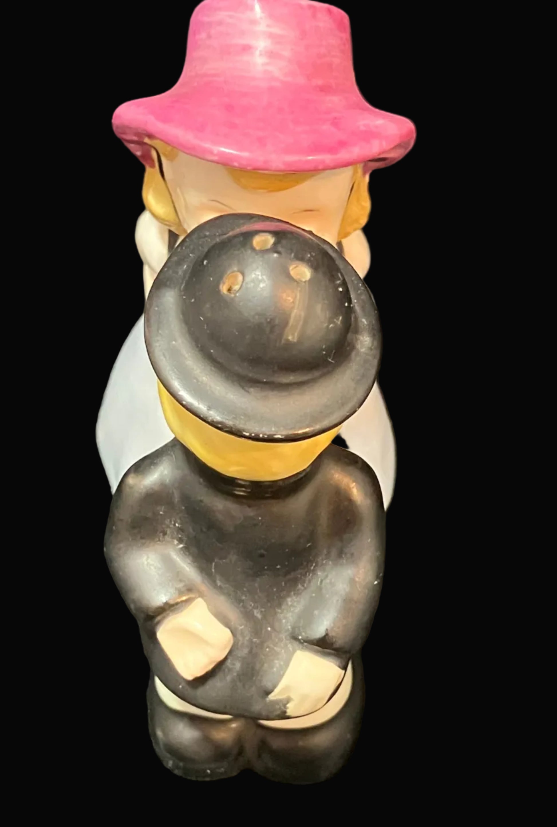 A vintage ceramic figurine depicting a couple, with the woman wearing a pink hat and the man in a dark gray suit and bowler hat, set against a solid black background.