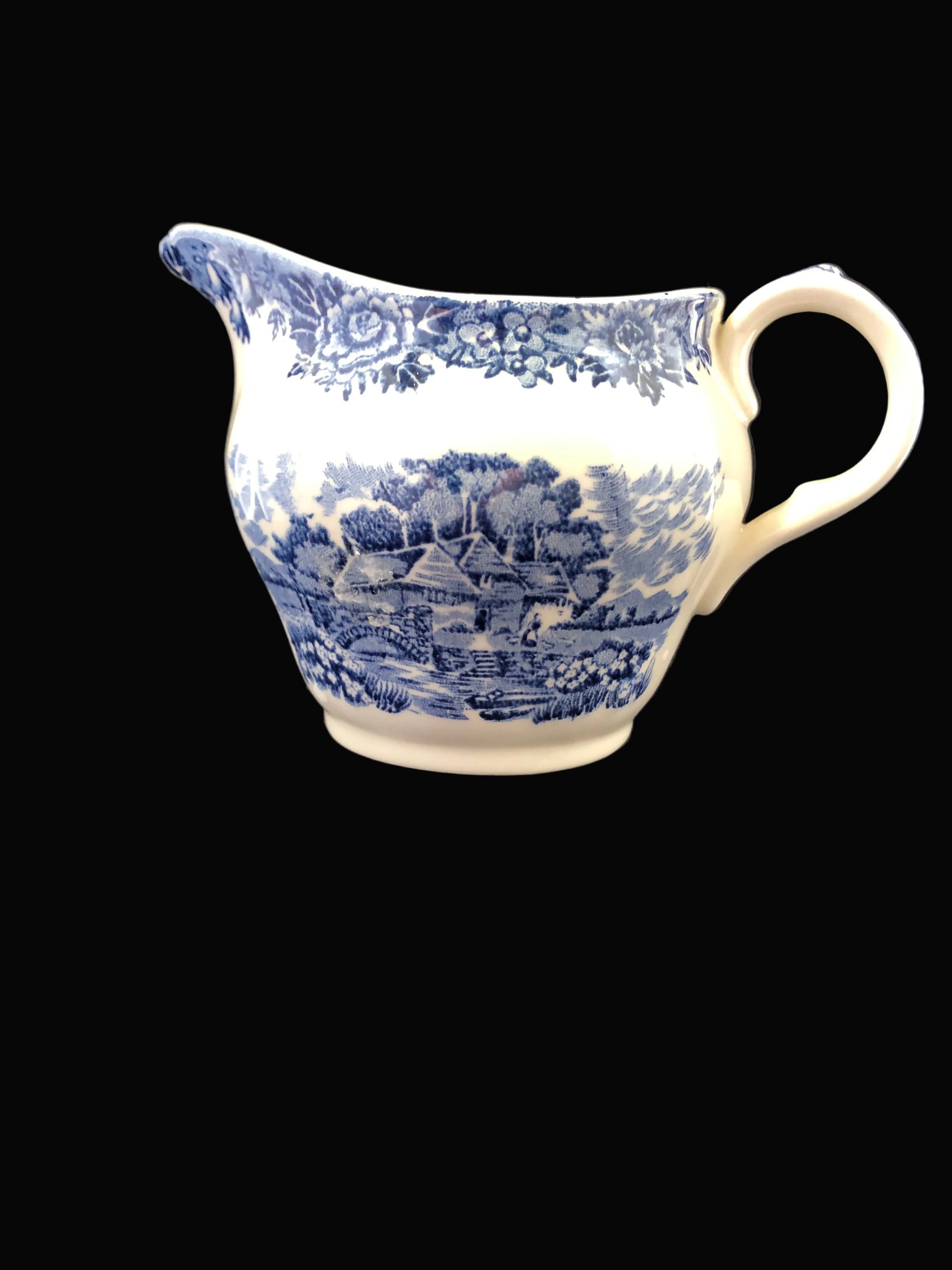 A white ceramic creamer pitcher with blue transfer landscape and floral patterns, set against a dark  background.