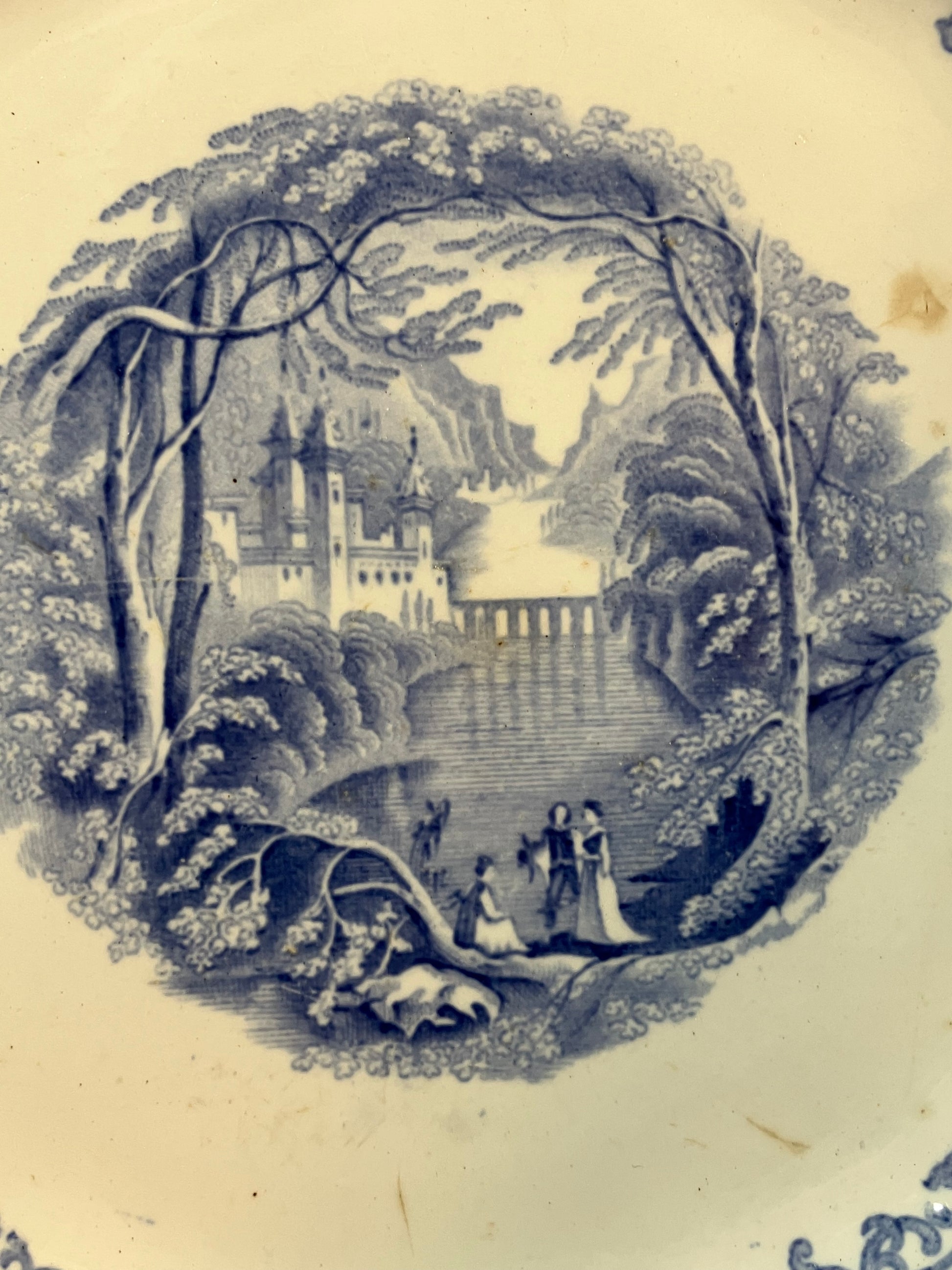 A close-up of a vintage ceramic plate or dish featuring a blue and white printed scenic illustration of a castle by a river, framed by trees, with small figures near the water in the foreground.