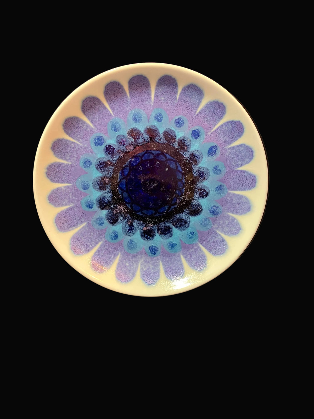 A round ceramic plate with a daisy-like floral pattern, featuring a cream-colored rim, a blue and purple center, and purple petals radiating outward, set against a dark background.