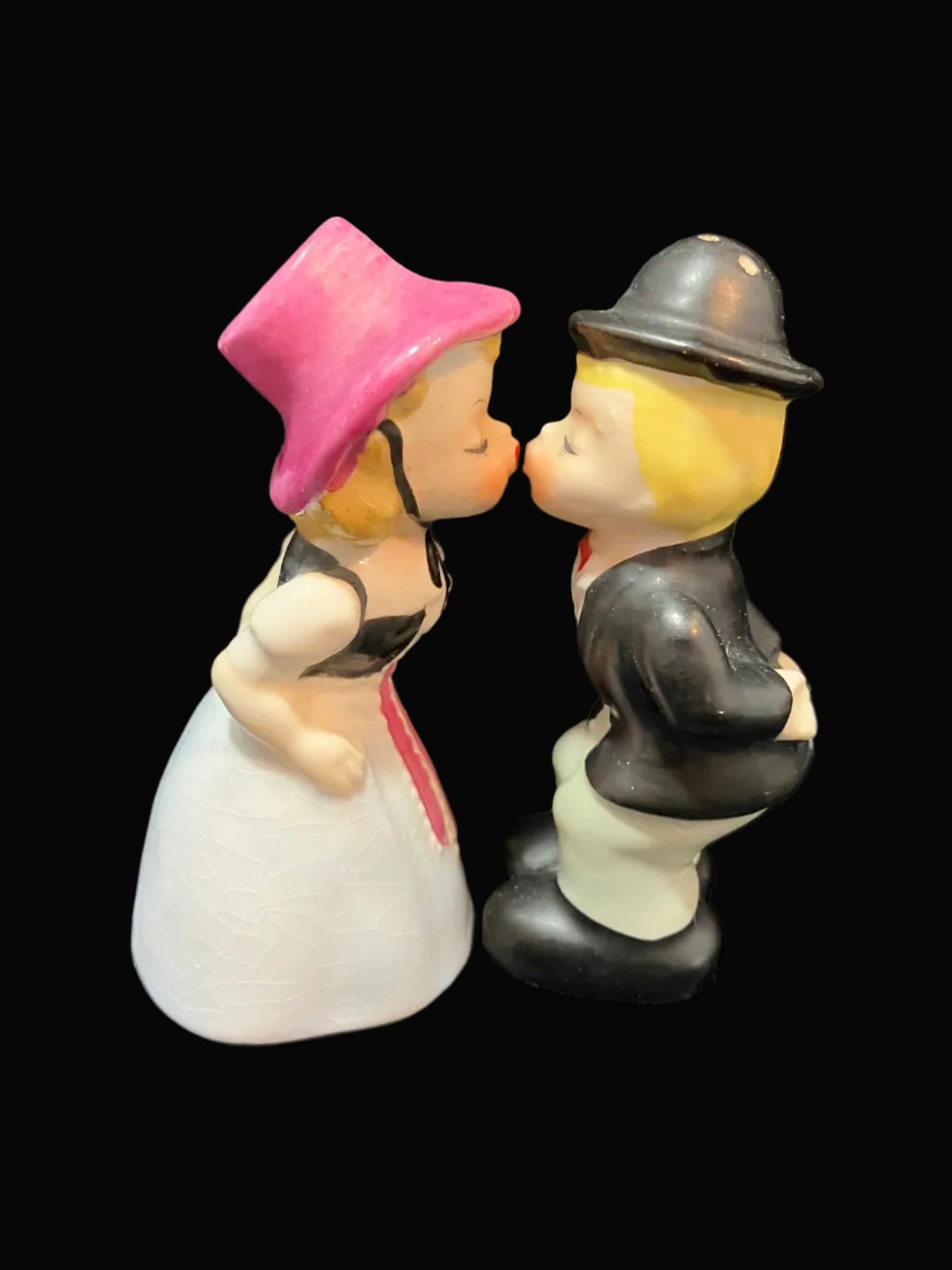 A vintage ceramic figurine of a kissing couple, featuring a woman in a pink hat and white dress, and a man in a black suit and bowler hat, set against a solid black background.