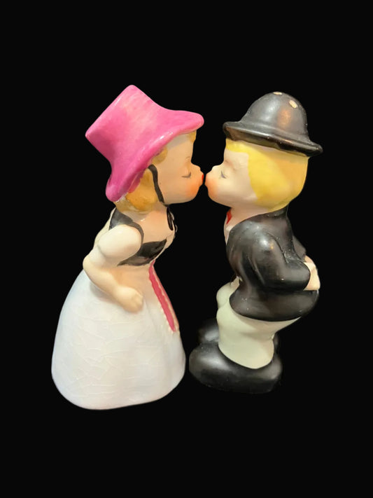 A vintage ceramic figurine of a kissing couple, featuring a woman in a pink hat and white dress, and a man in a black suit and bowler hat, set against a solid black background.
